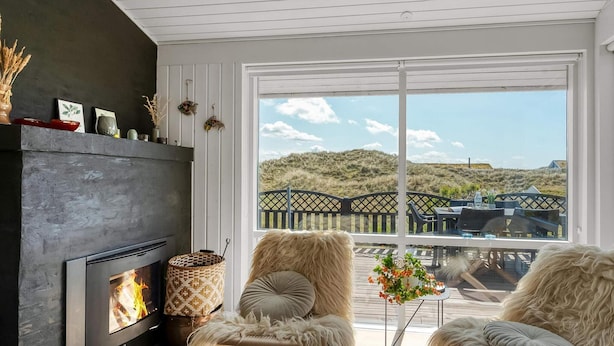 A cozy room with a fireplace, two plush chairs, a small table with flowers, and a large window overlooking a scenic landscape. - 4 Star Holiday Home in Saltum (Saltum)