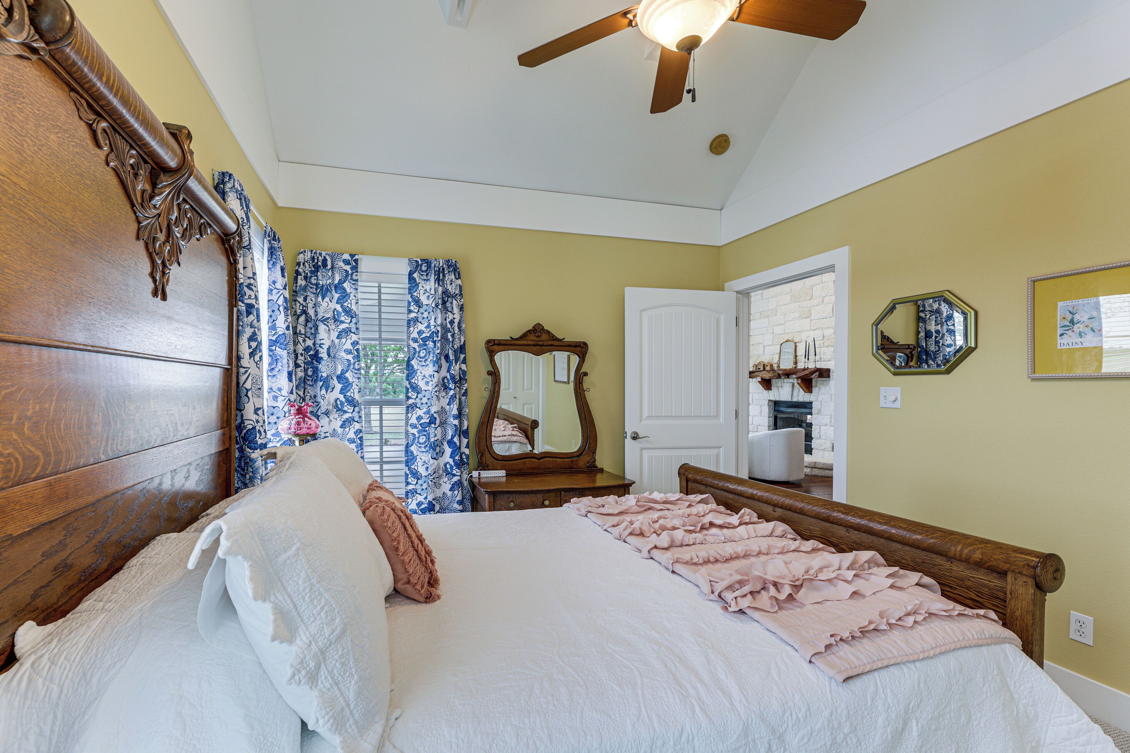 Quarto com cabeceira de madeira, ventilador de teto, cama com roupa de cama branca e rosa, cômoda, espelho e porta que dá para outro ambiente.