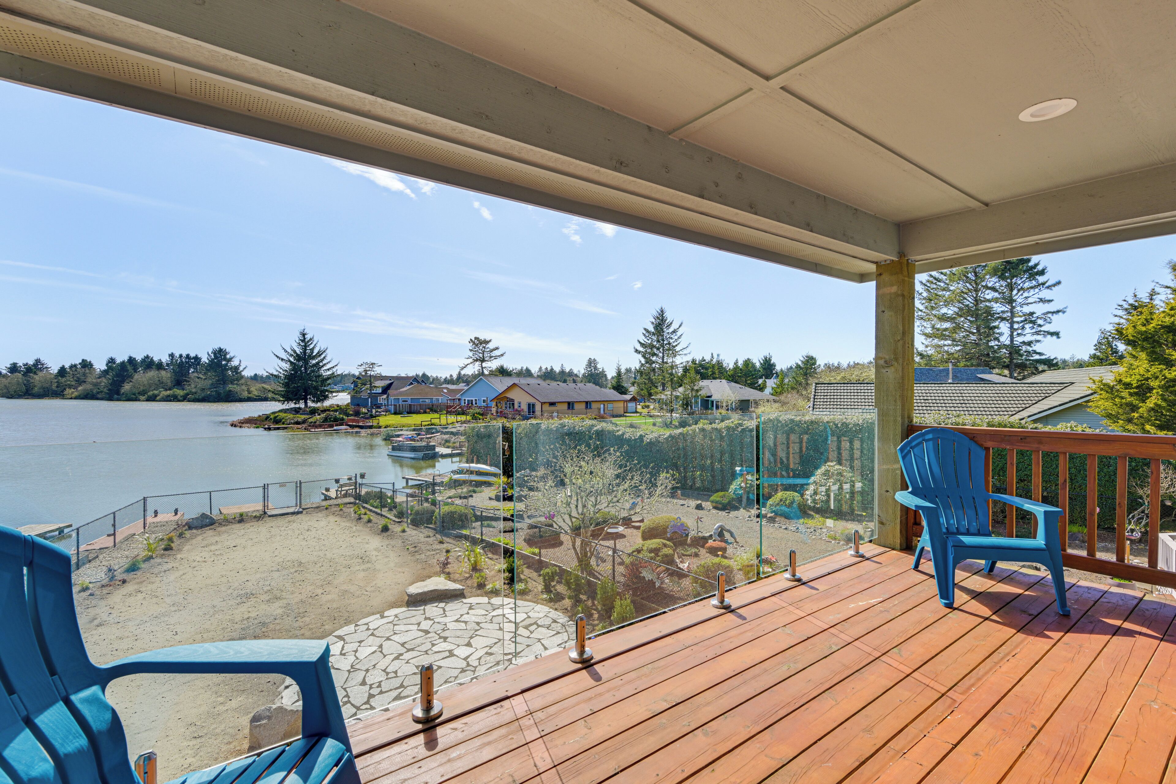 Una terraza de madera con dos sillas azules, con vistas a un lago y casas y árboles al fondo.