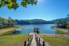 Une jetée en bois mène à un petit bateau amarré et à un chalet situés sur un lac paisible, entouré de collines boisées.