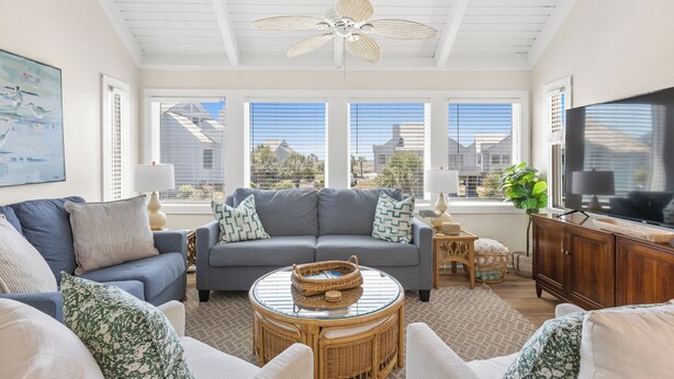 A living room with a grey sofa, a wicker coffee table, and a television on a wooden cabinet. Large windows provide views of buildings and palm trees. - Your seat for sweeping ocean views and unforgettable sunsets at Villa 22 (Bald Head Island)