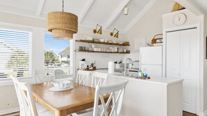 A bright kitchen with white cabinetry, a wooden dining table, and white chairs. There are hanging pendant lights, a white refrigerator, and a clock on the wall. - Your seat for sweeping ocean views and unforgettable sunsets at Villa 22 (Bald Head Island)