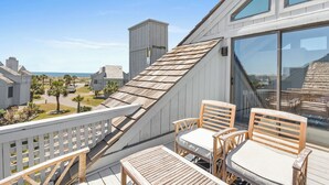 A balcony with wooden furniture, a view of the beach, and a building with large windows. - Your seat for sweeping ocean views and unforgettable sunsets at Villa 22 (Bald Head Island)