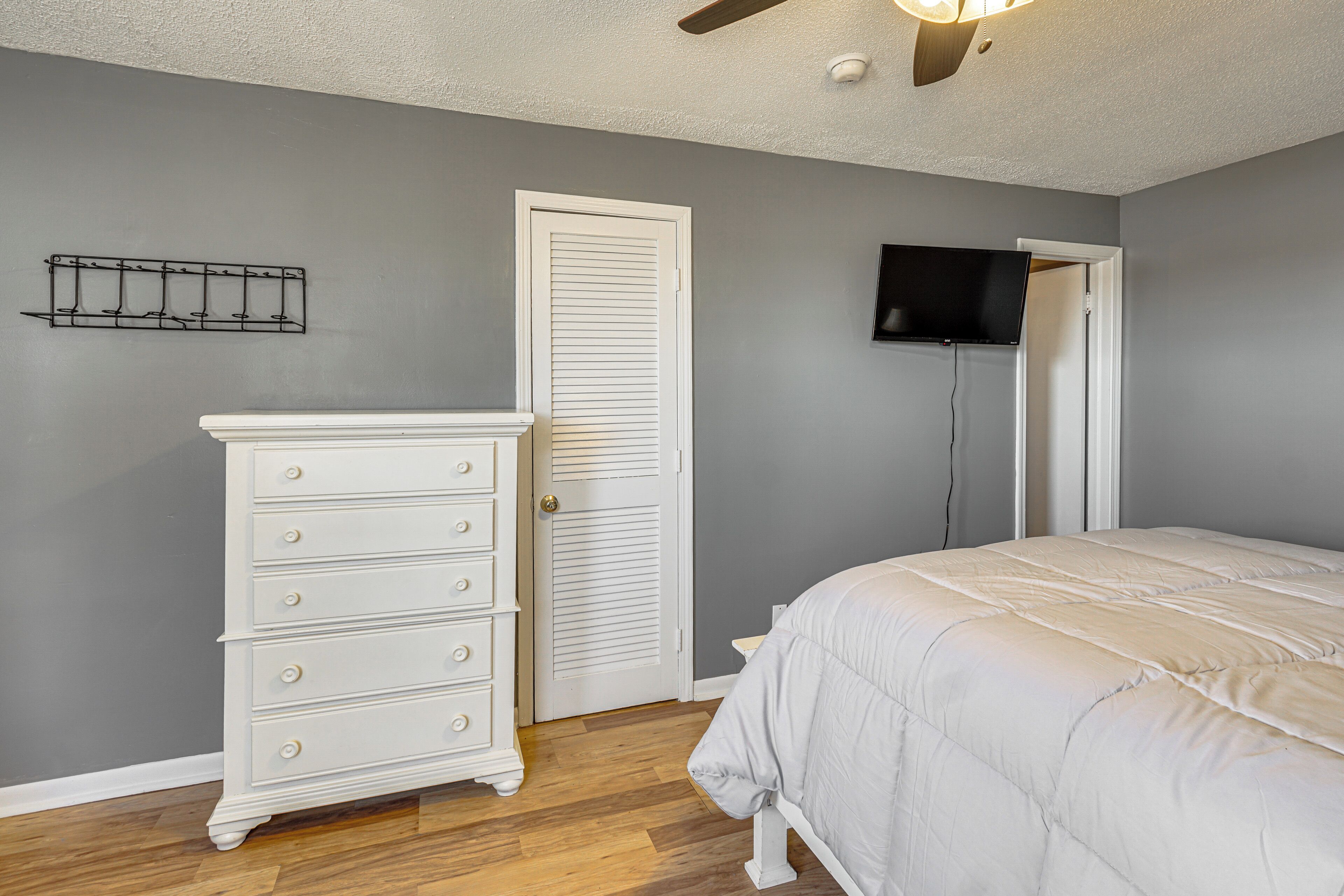 A bedroom with a white dresser, a bed with a white comforter, a ceiling fan, a mounted TV, and a door with a white frame.