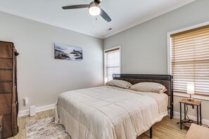 A bedroom with a bed, a ceiling fan, a nightstand with a lamp, a painting on the wall, and a window with blinds.
