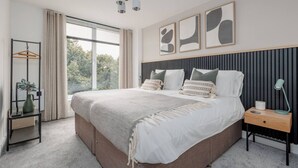Une chambre moderne avec un grand lit, une table de chevet avec une lampe, une plante suspendue et une vue sur l’extérieur par une fenêtre.