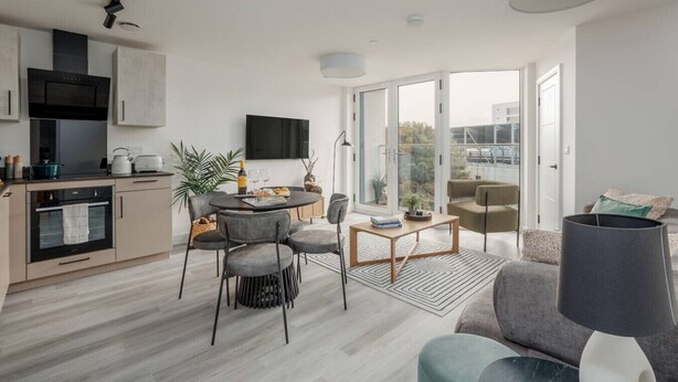Un appartement moderne comprenant une cuisine, un coin repas et un salon. Il est équipé d’un téléviseur à écran plat, d’une table à manger avec des chaises, d’un canapé et d’une grande fenêtre offrant une vue sur la ville.