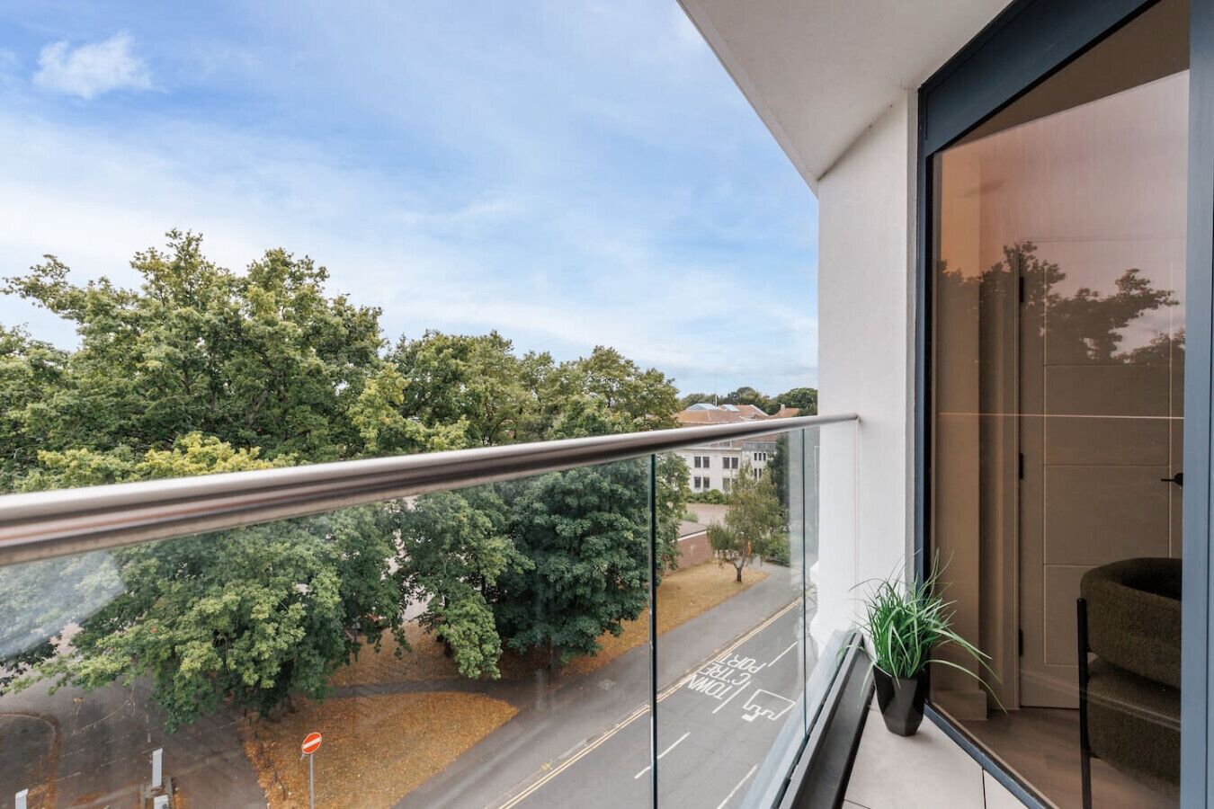 A balcony with a glass railing overlooking a road and trees, a potted plant, and a view of a building across the street.