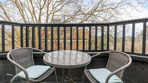 Ein Balkon mit zwei Korbstühlen und einem runden Tisch, mit Blick auf ein Backsteingebäude und kahl stehende Bäume.