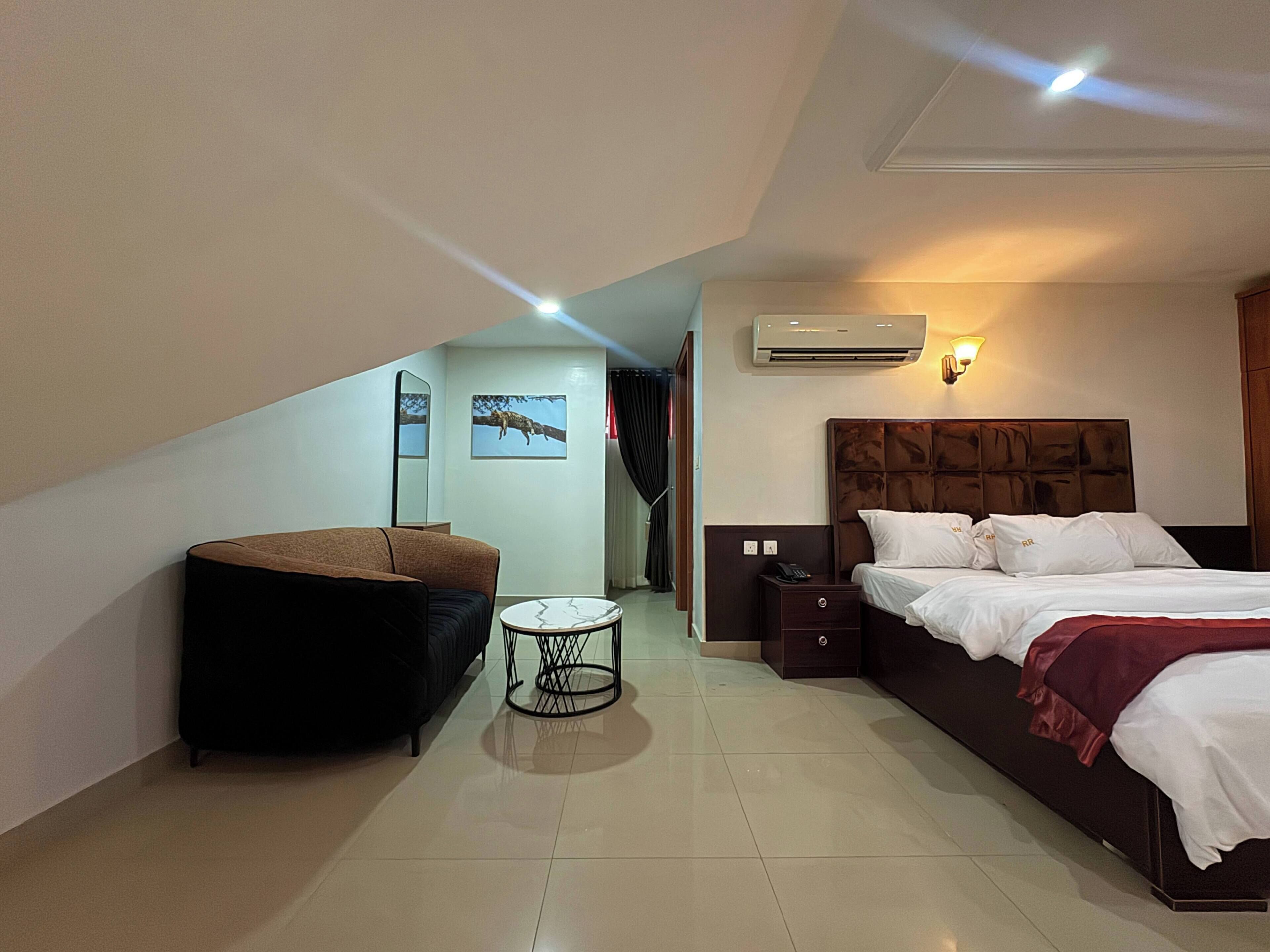A hotel room with a bed, a chair, a small table, an air conditioner, and a mirror.