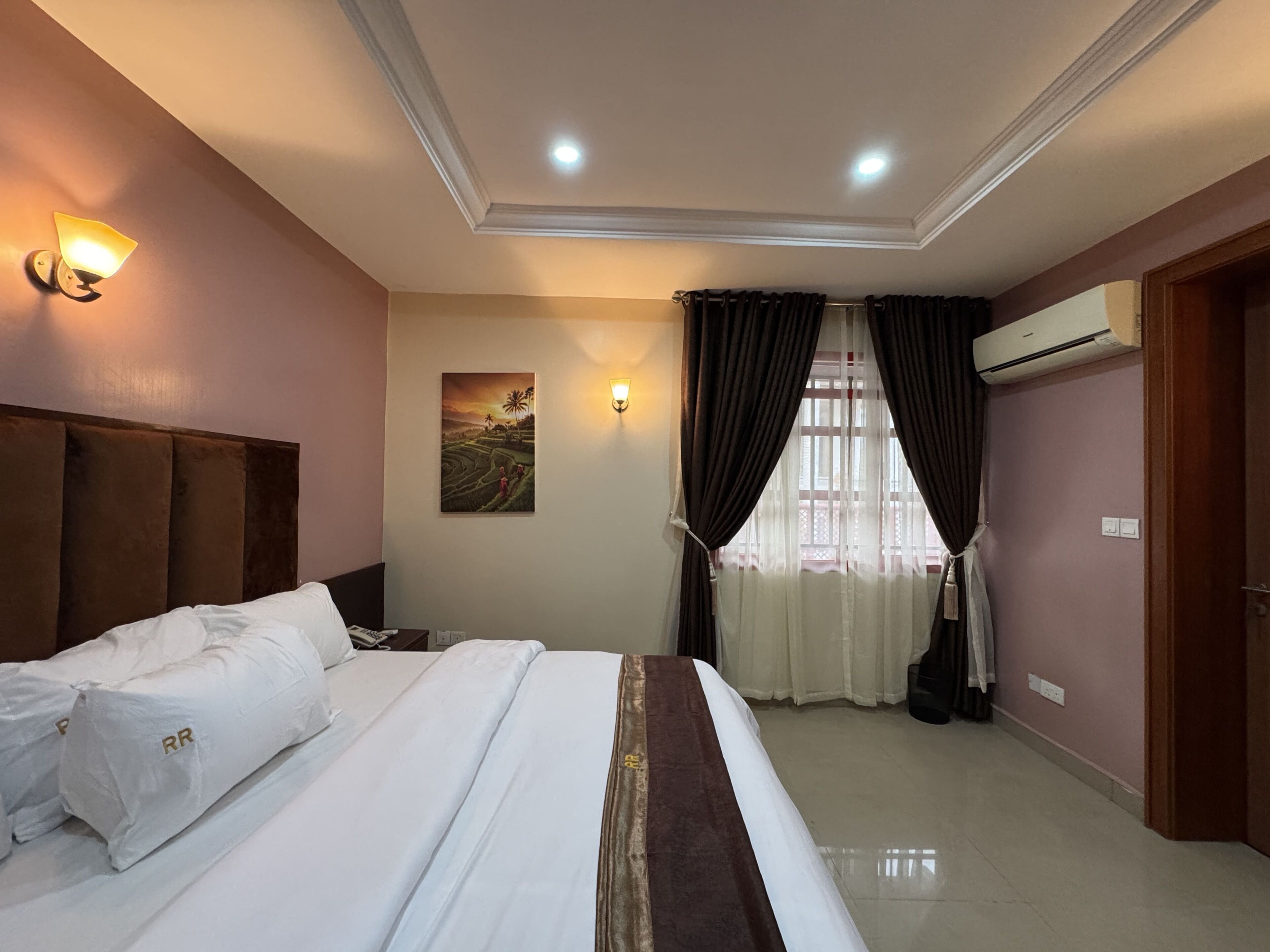 A hotel room with a bed, a wall-mounted light, an air conditioning unit, and a painting on the wall.