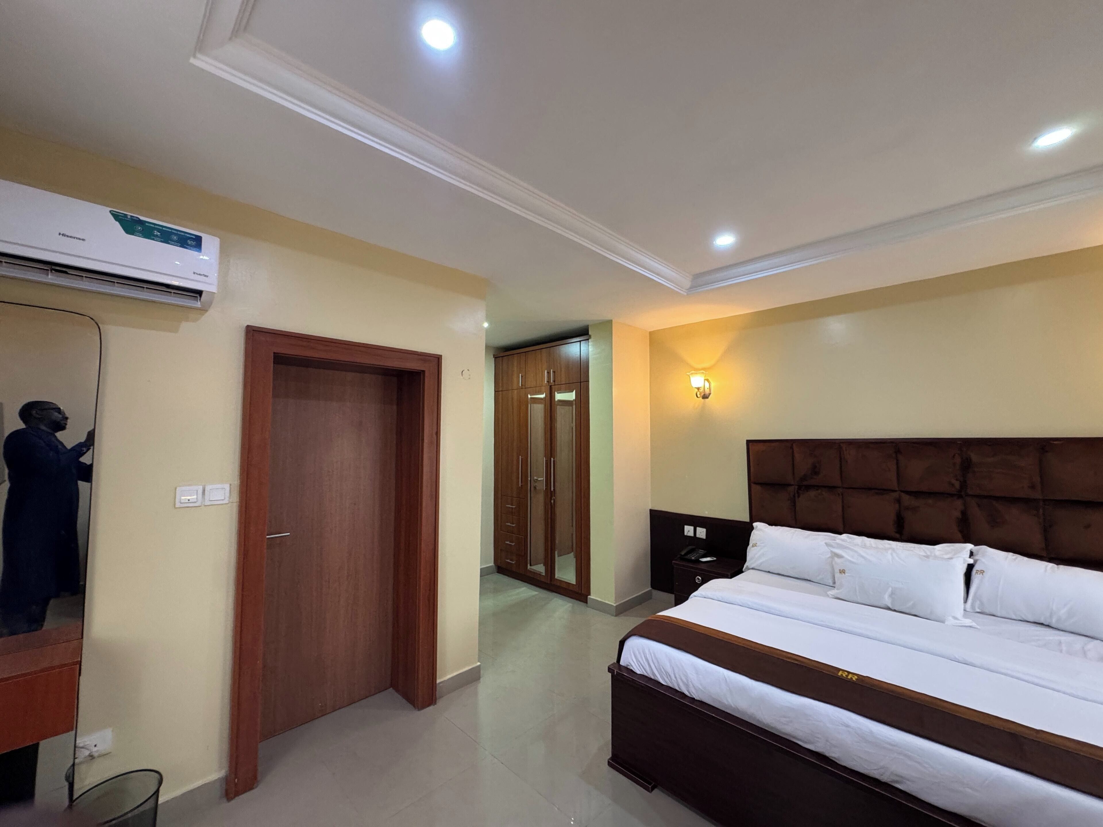 A hotel room with a bed, a wardrobe, a mirror, an air conditioning unit, and a nightstand.