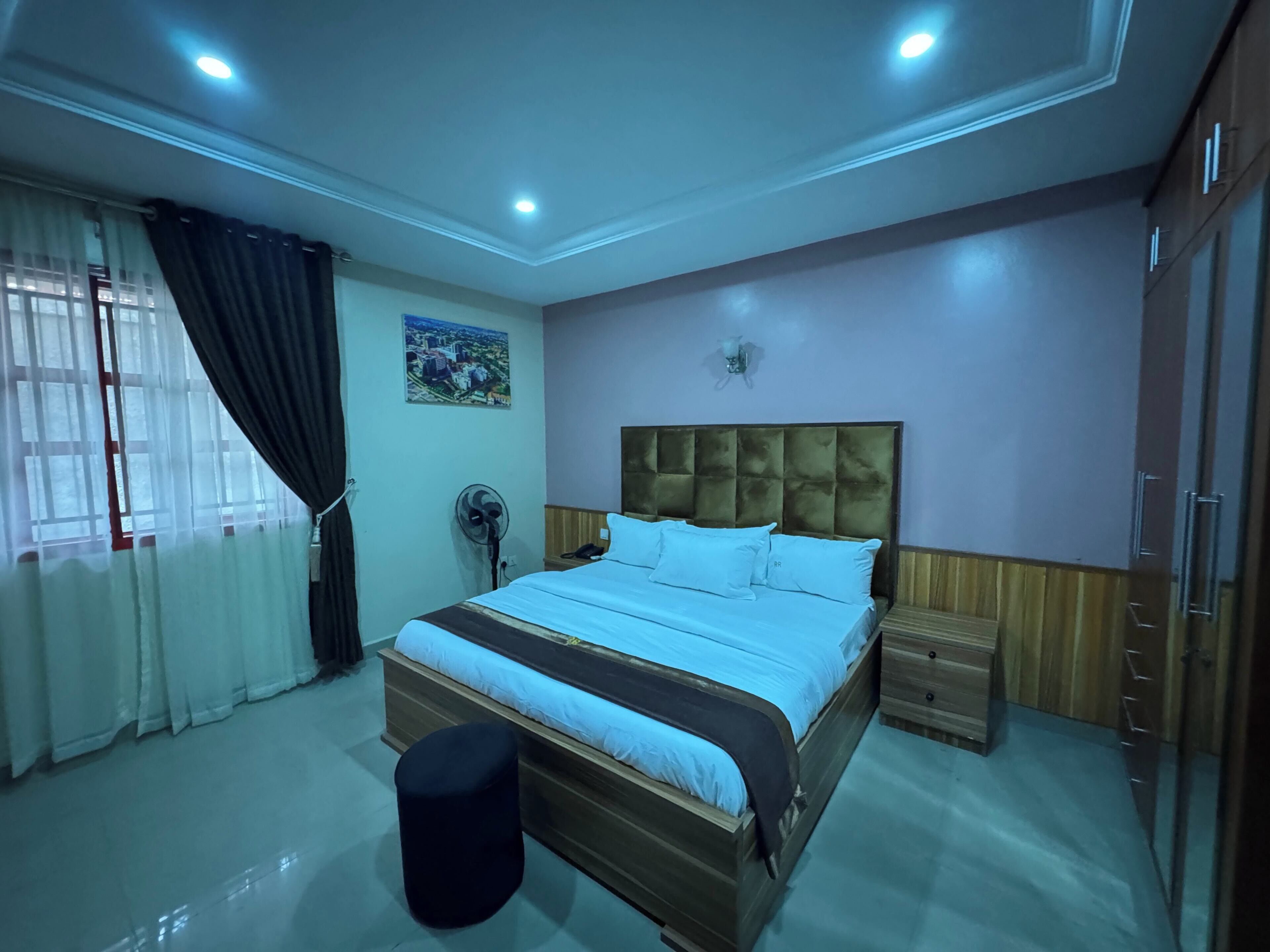 A bedroom with a bed, bedside tables, a fan, a nightstand, a wall-mounted light, and a window with curtains.