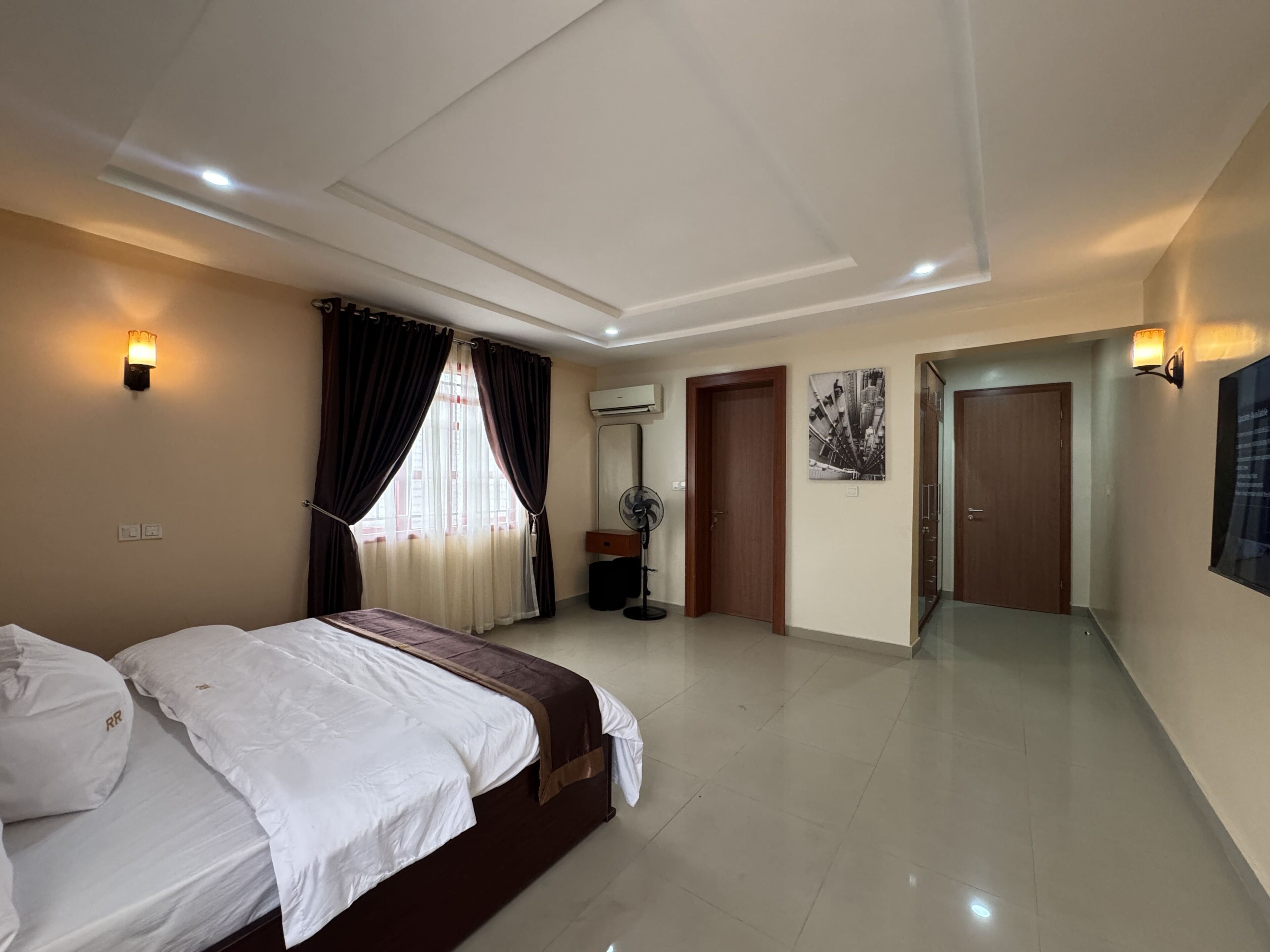 A hotel room with a bed, a standing fan, an air conditioning unit, a door, and a wall-mounted light fixture.