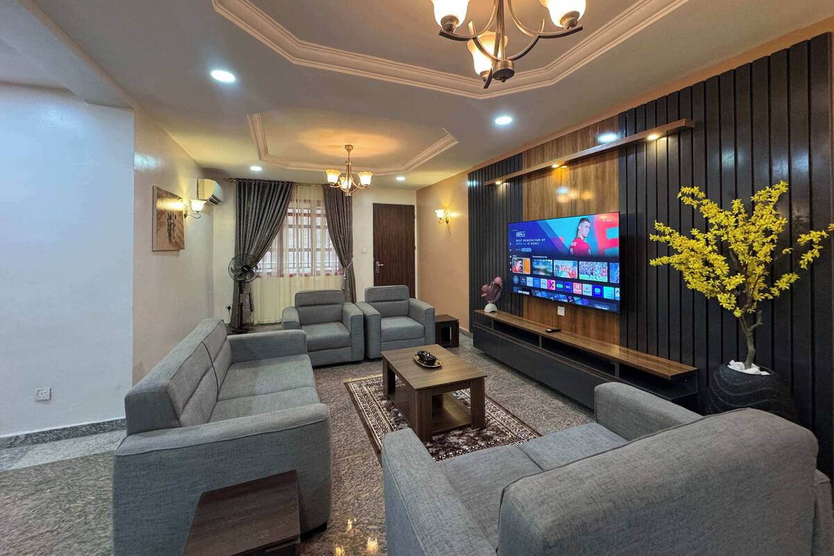A living room with a flat-screen TV, grey sofas, a coffee table, a chandelier, and a decorative plant.