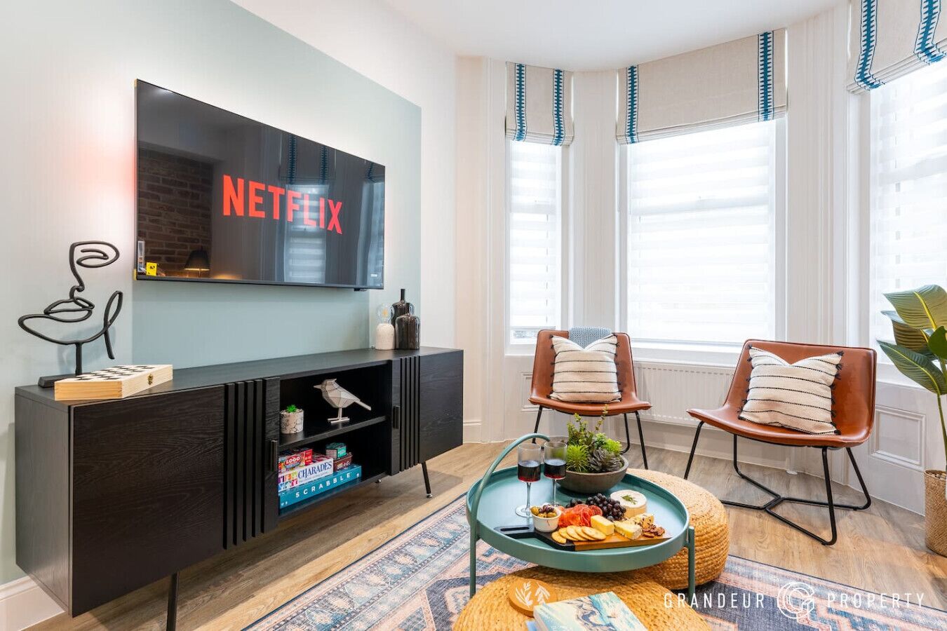 A modern living room with a flat-screen TV, a black entertainment unit, two leather chairs, a coffee table with snacks, and a large window with blinds.