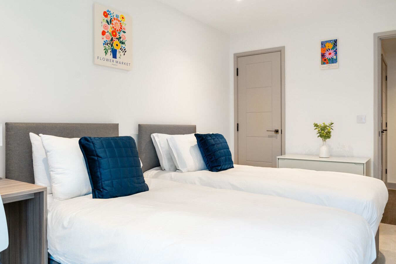 A hotel room with two beds, a bedside table, a nightstand, a door, and a small white cabinet.