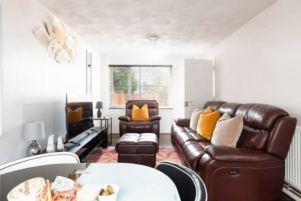 Un salon avec un canapé en cuir marron, un fauteuil en cuir marron, une table à manger recouverte d’une nappe blanche et un téléviseur.