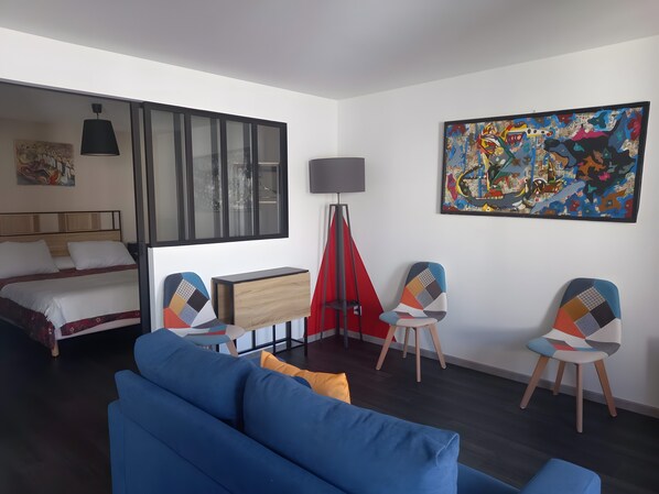A modern hotel room with a bed, blue sofa, two chairs, a desk, a lamp, and a large abstract painting.