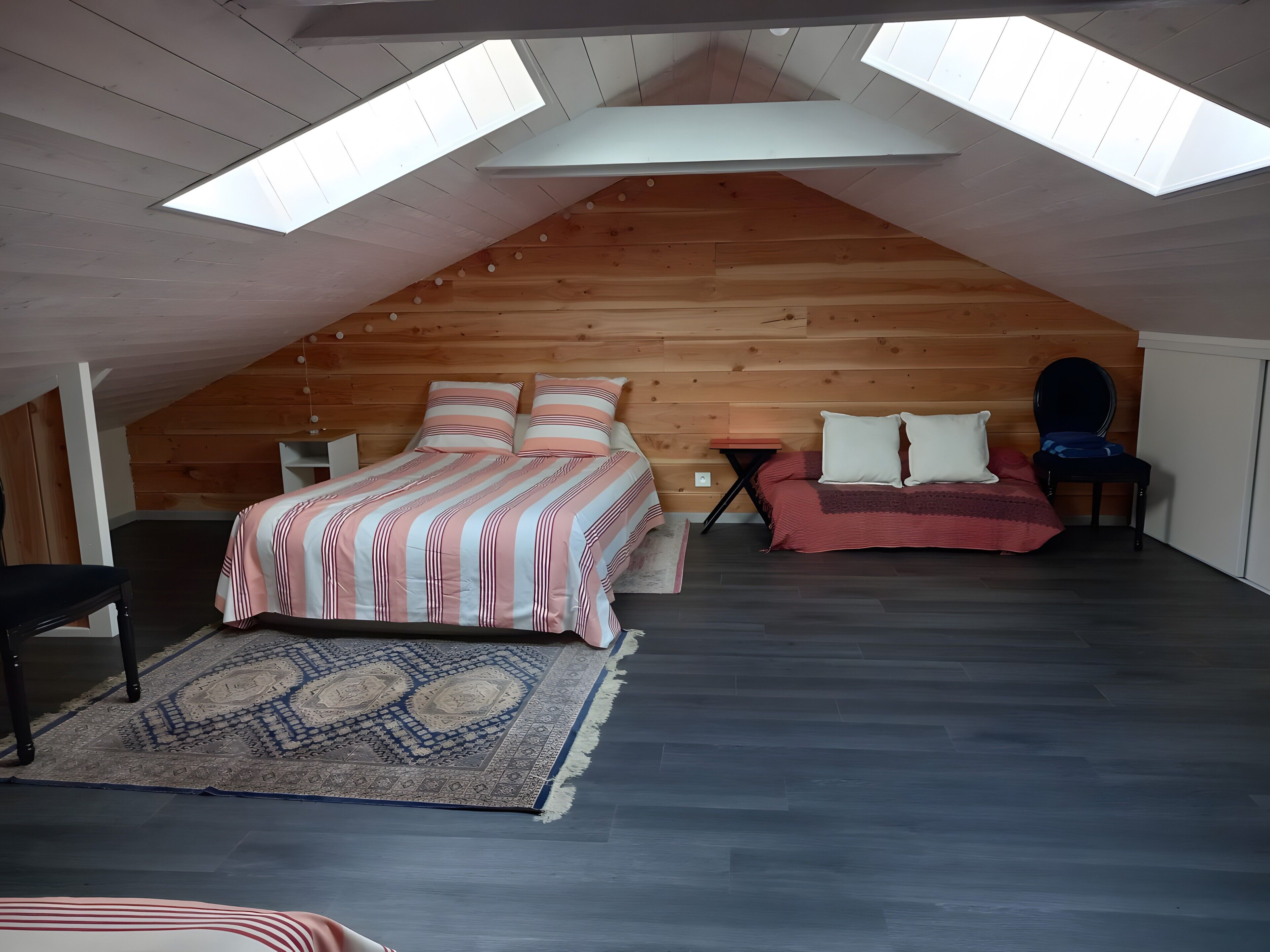 Une chambre à coucher aménagée dans un espace sous les toits, avec un plafond en pente, des murs en bois, un lit avec une literie à rayures, un canapé rouge et un tapis à motifs.