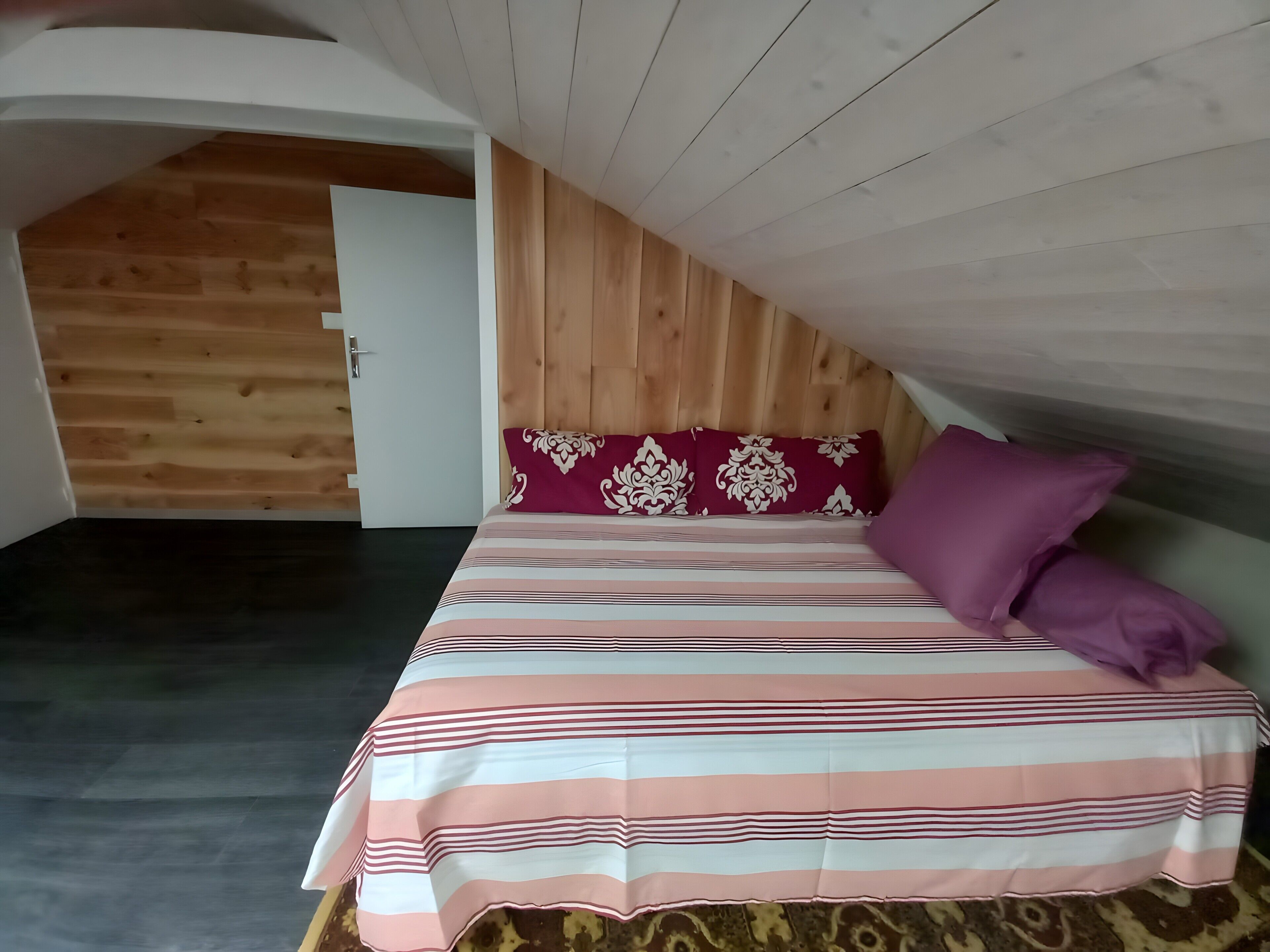 Un dormitorio con techo inclinado, una cama con ropa de cama a rayas, un cabecero de madera y una puerta.