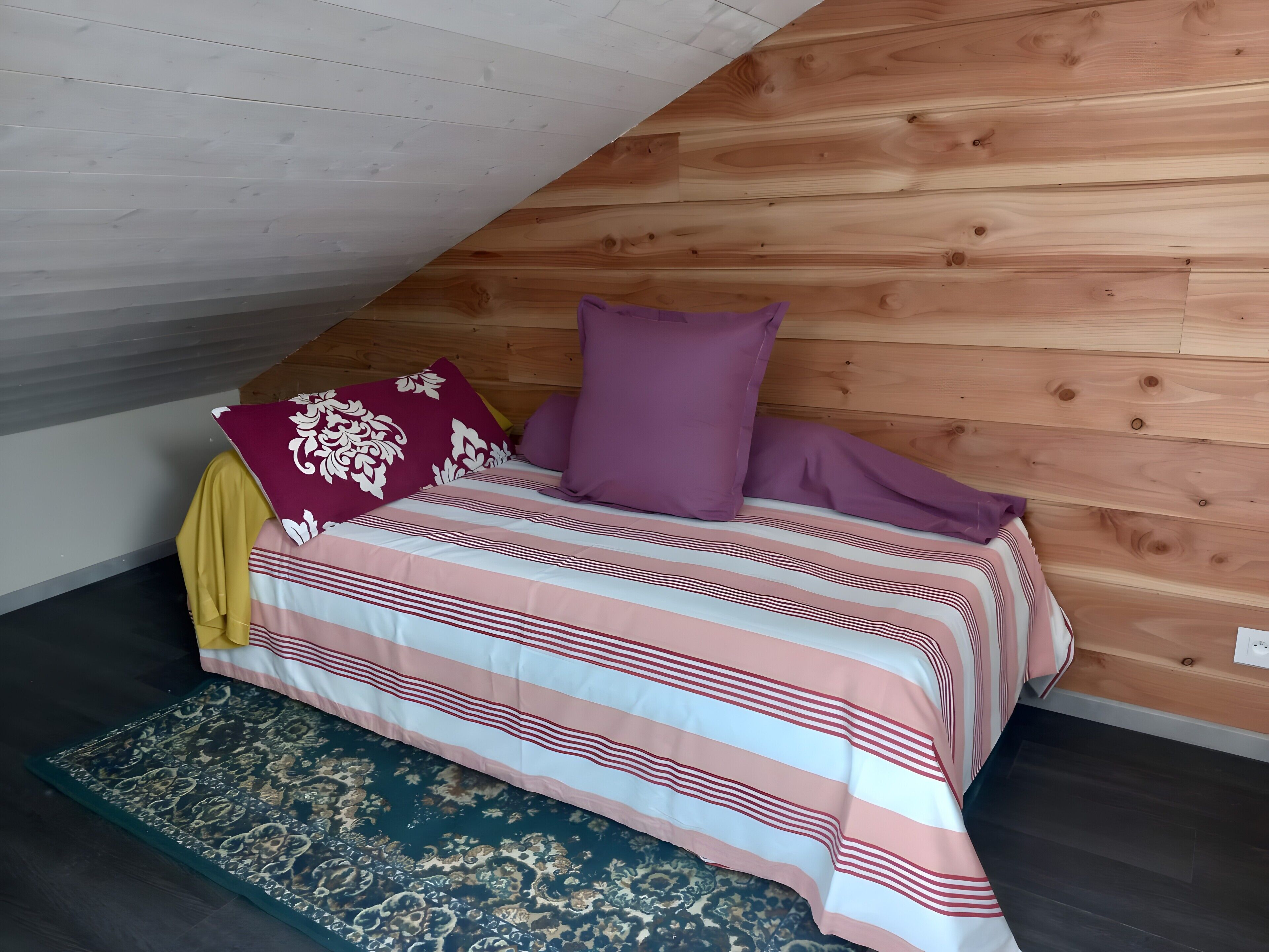 Une chambre avec un plafond en pente, des murs recouverts de lambris en bois, un lit avec une literie à rayures et un tapis à motifs.