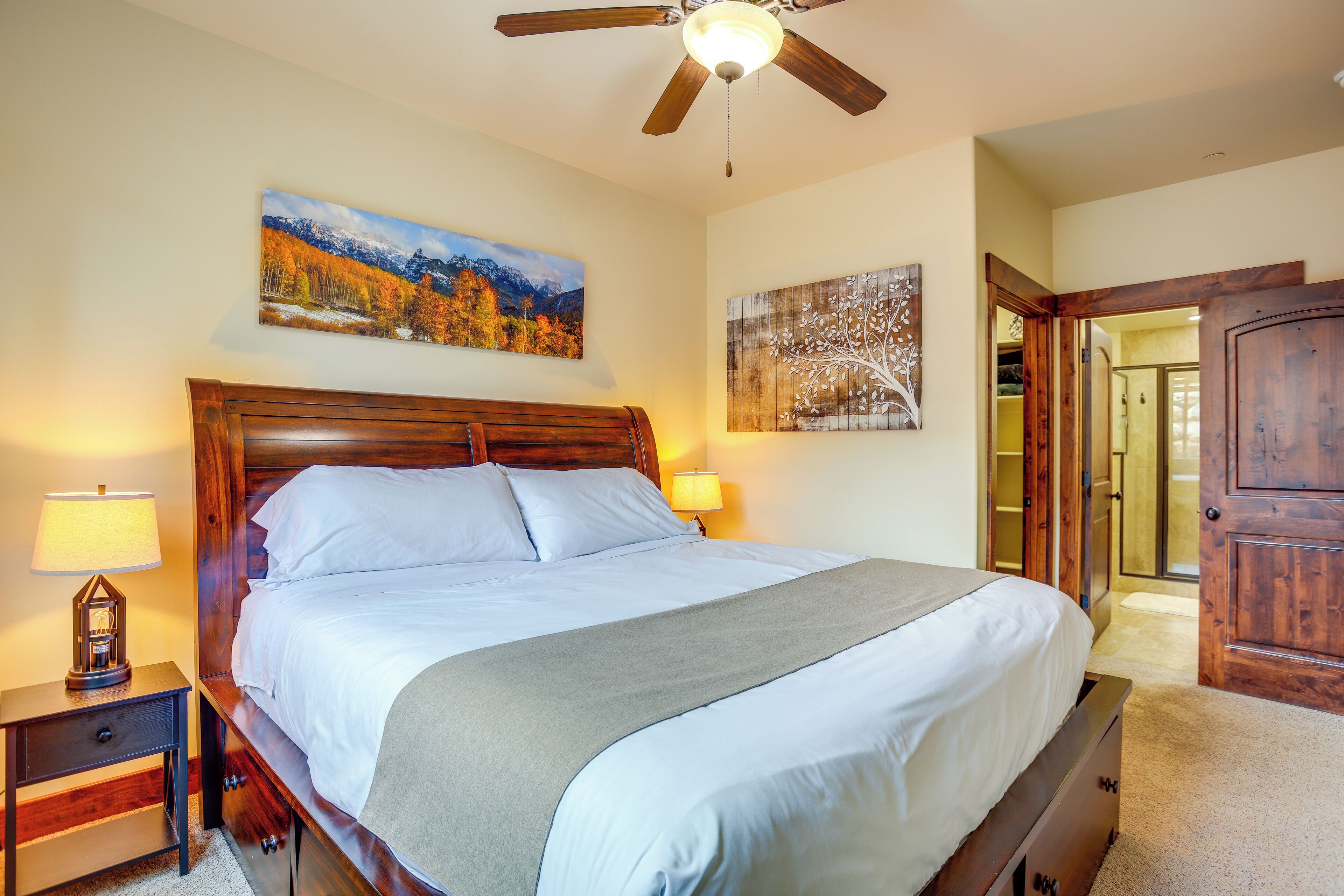 A bedroom with a wooden bed, two bedside tables with lamps, a ceiling fan, and a painting on the wall.