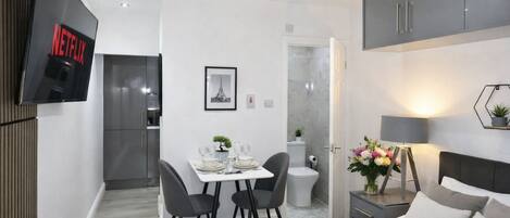 A modern hotel room with a bed, a TV mounted on the wall, a dining area with a table and chairs, a small bathroom, and a closet.