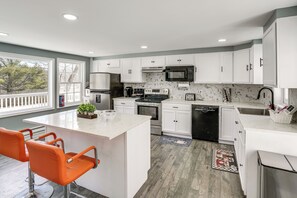 A modern kitchen with white cabinets, a central island, orange chairs, a large window, and a view of trees. - Indoor Hot Tub & Sauna! Spacious Catskills Home (Cairo)