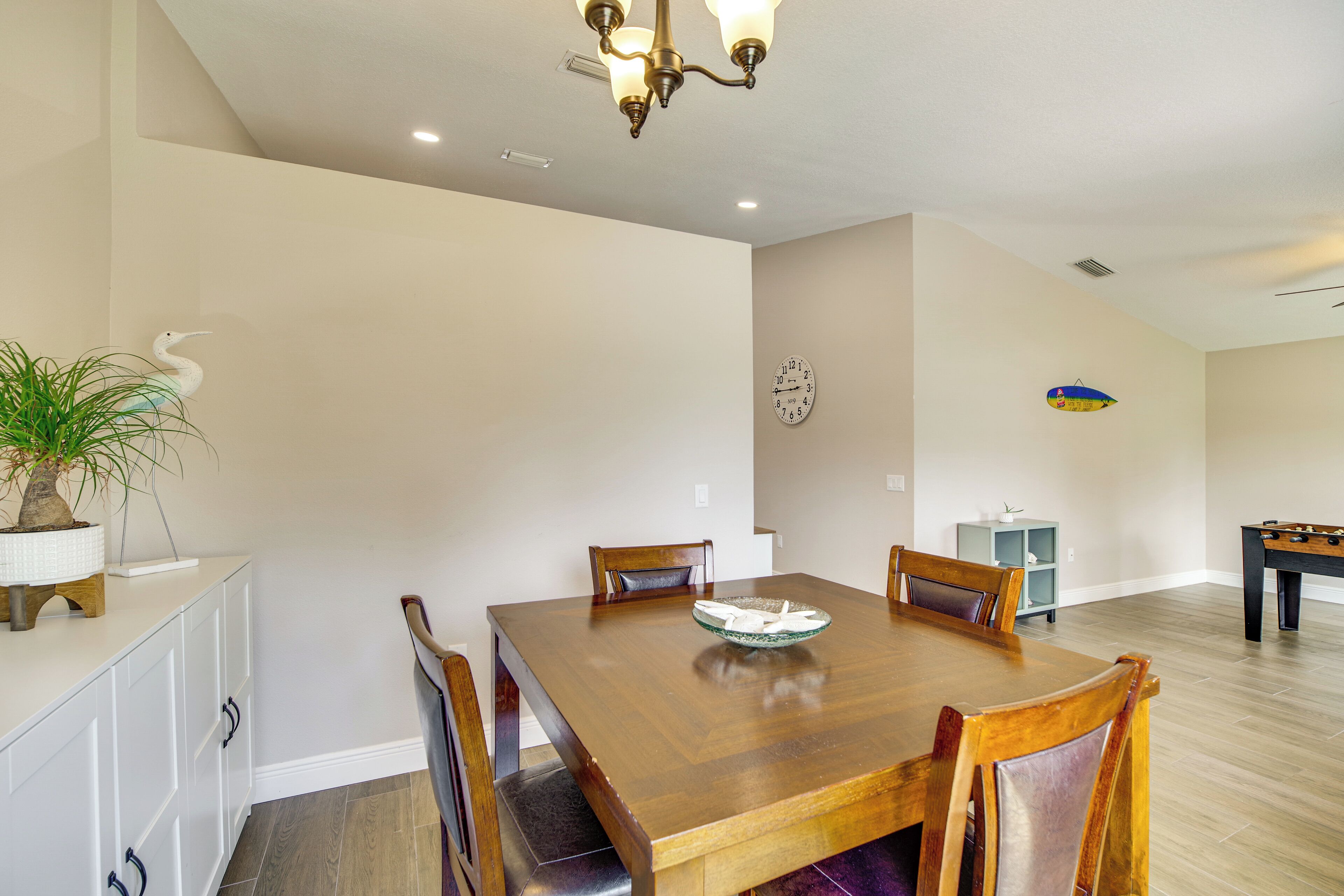 A dining area with a wooden table, chairs, a potted plant, a clock, and a foosball table.