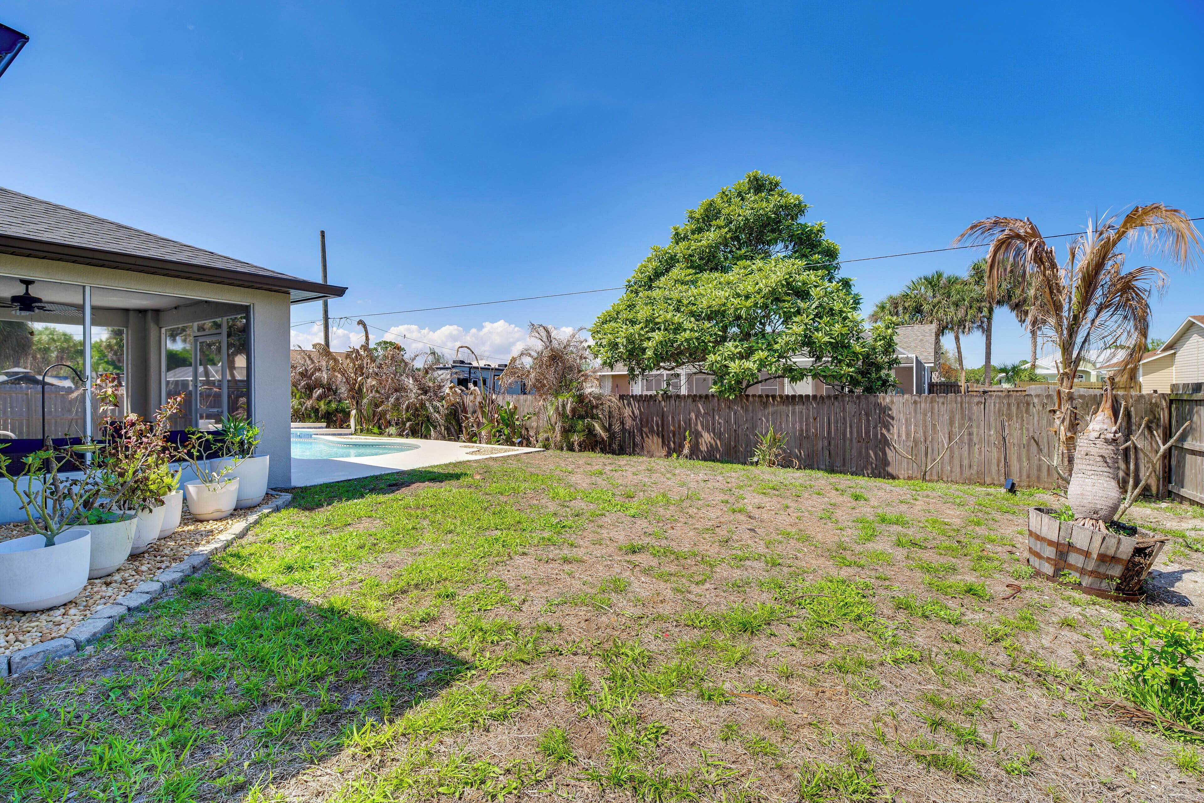 A backyard with a swimming pool, potted plants, a wooden fence, and a large tree.