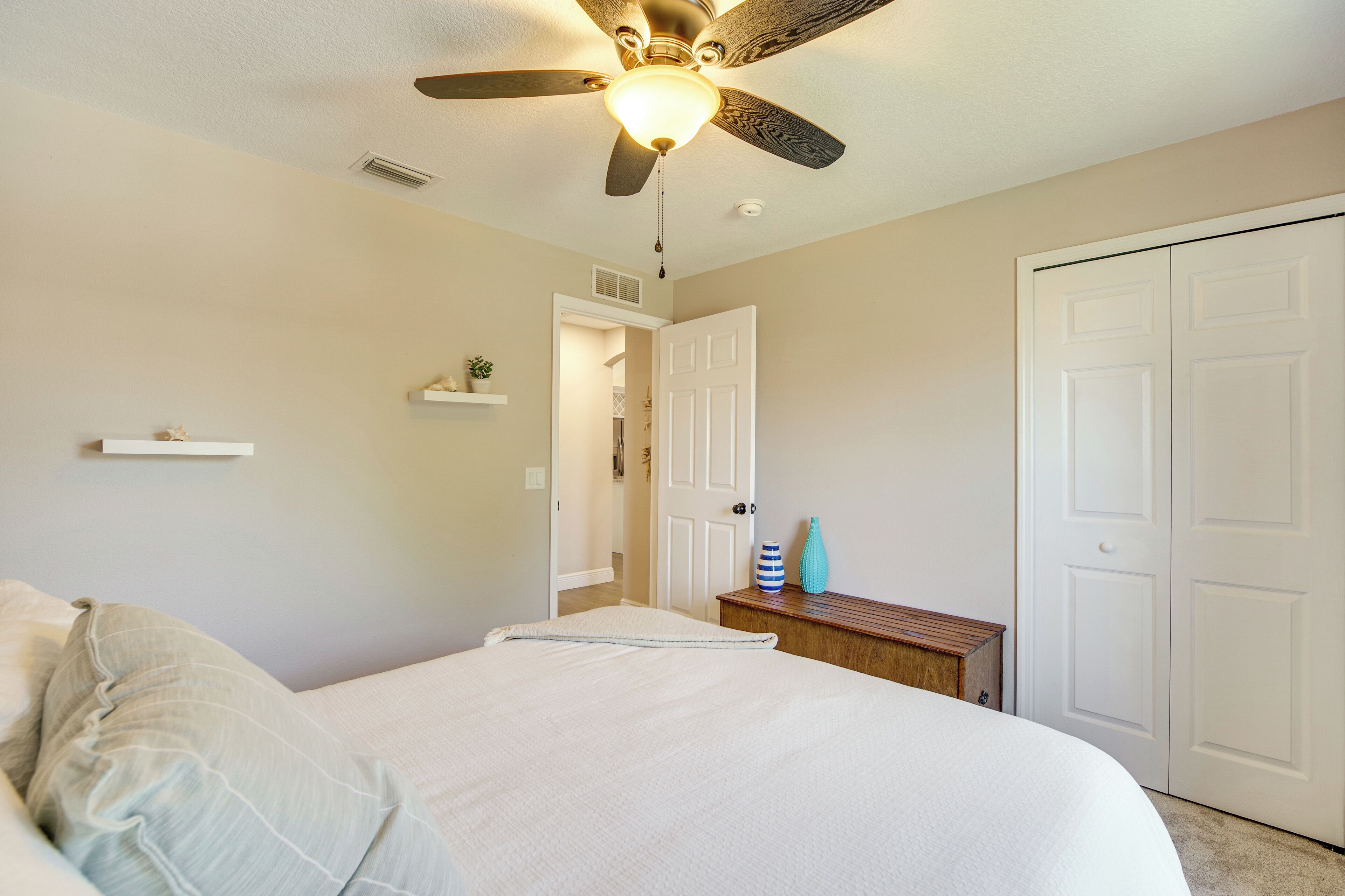 Un dormitorio con cama, ventilador de techo, puerta y una pequeña mesa con adornos.