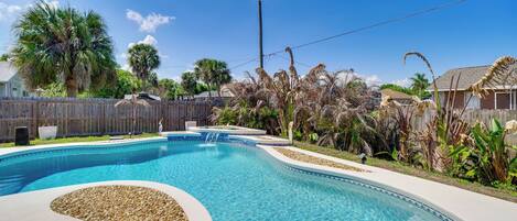 A swimming pool with a pebble design, surrounded by a wooden fence and palm trees.