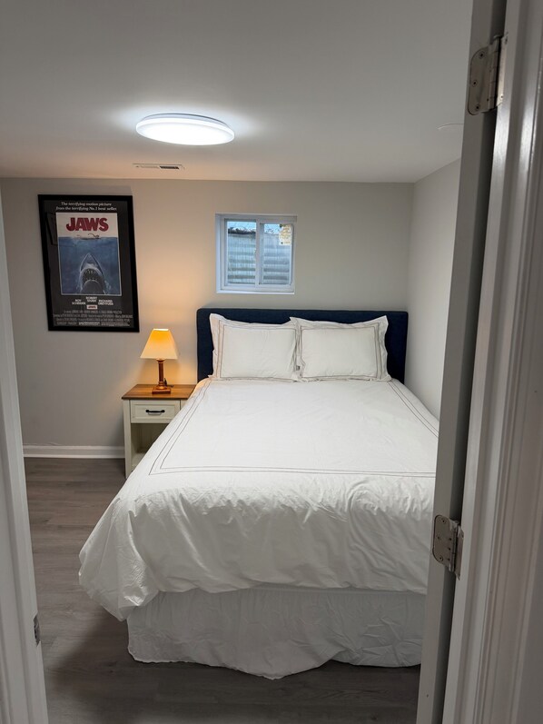 Une chambre à coucher avec un lit, une table de chevet avec une lampe, une affiche au mur et une fenêtre.
