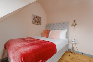 A bedroom with a bed, a red blanket, a white mattress, a grey headboard, a small bedside table with a clock, and a floor lamp.