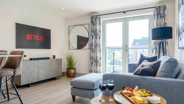 Ein modernes Wohnzimmer mit einem grauen Sofa, einem Essbereich mit Tisch und Stühlen, einem großen Fenster mit Vorhängen und einem an der Wand befestigten Netflix-Schild.