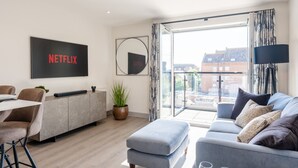 Ein modernes Wohnzimmer mit einer Couch, einem Couchtisch, einem an der Wand befestigten Fernseher, einer Pflanze und Blick auf die Stadt durch große Fenster.