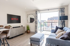 A modern living room with a sofa, a coffee table, a TV mounted on the wall, a plant, and a view of the city through large windows. - Quay View - Close to the quay & town with balcony & parking (Poole)