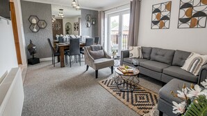 A modern living room with a grey sofa, a dining area with a wooden table, and a large window with curtains. - The Catalina Breeze - By the harbour, park and town with parking (Dorset)