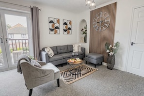 A modern living room with a grey sofa, a coffee table with a fruit platter, a grey armchair, a patterned rug, and a large wall clock.
