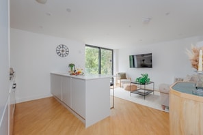 A modern kitchen with white cabinetry, a large window, a flat-screen TV, and a dining area with a glass table and chairs.