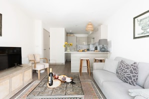 Un salon moderne avec un canapé, une table basse sur laquelle se trouvent une bouteille et des verres, un téléviseur, et une cuisine en arrière-plan.