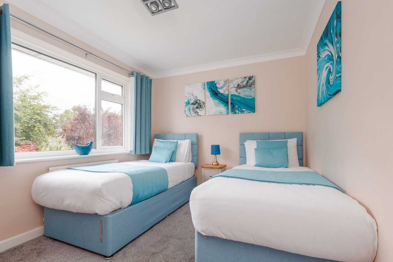 A bedroom with two beds, a window with curtains, a nightstand with a lamp, and artwork on the wall.