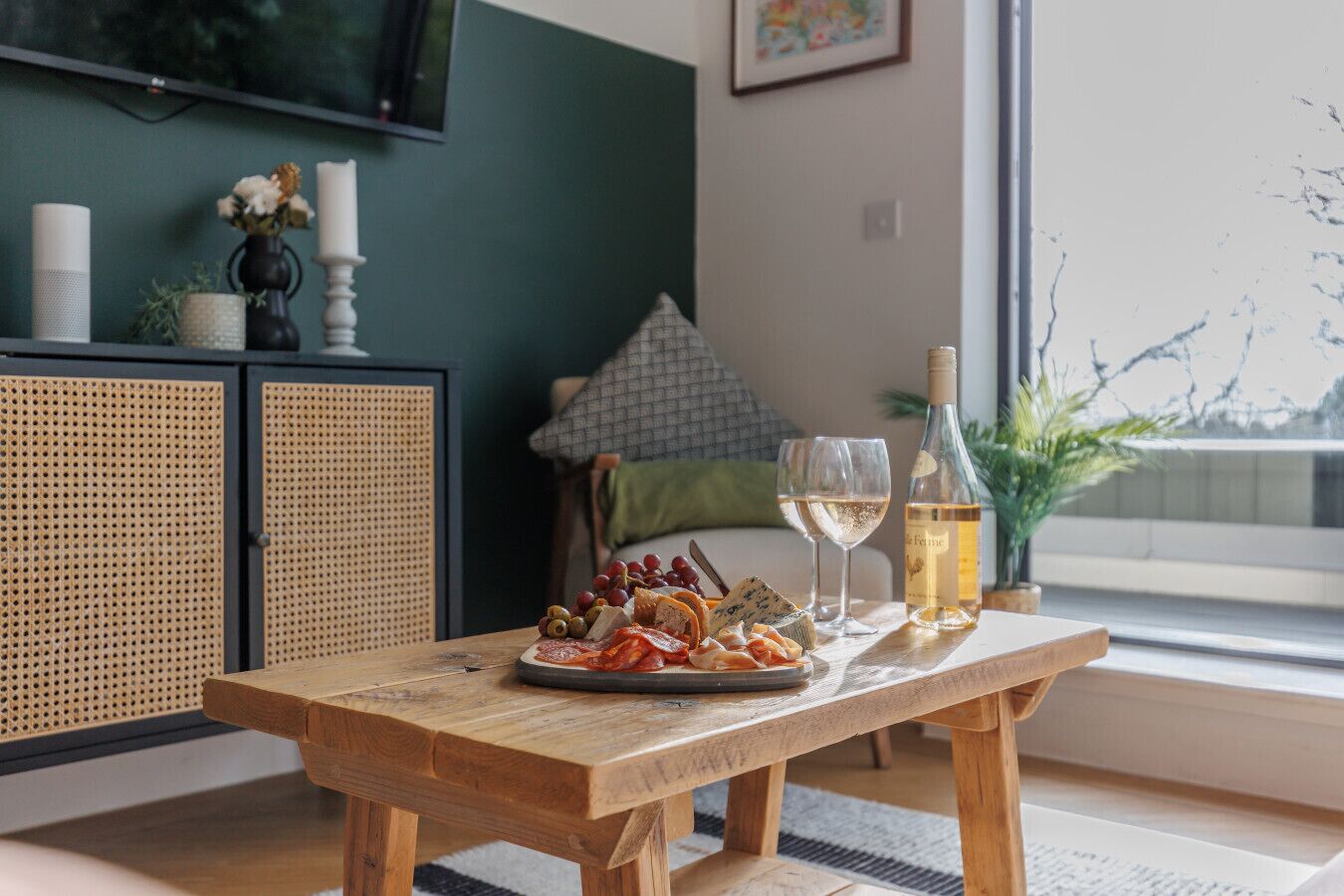 A wooden table with a charcuterie board, wine glasses, and a bottle of wine in a room with a large window, a television, and a green wall.