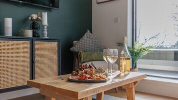 A wooden table with a charcuterie board, wine glasses, and a bottle of wine in a room with a large window, a television, and a green wall.