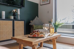 Ein Holztisch mit einer Wurstplatte, Weingläsern und einer Weinflasche in einem Raum mit großem Fenster, Fernseher und grüner Wand.