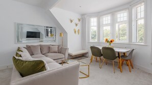 A modern living room with a sectional sofa, a dining table with chairs, a large window with blinds, and a ceiling with a sloped design. - Park Views - Great for places to eat and getting to the beach (Poole)
