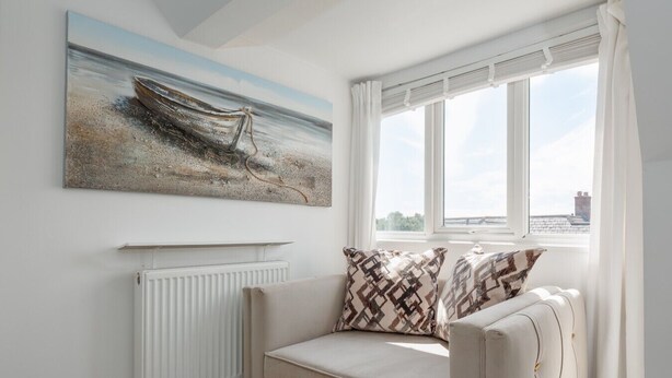 A room with a white radiator, a beige sofa with patterned cushions, a large painting of a boat on the wall, and a window with a view of the sky and buildings. - Park Views - Great for places to eat and getting to the beach (Poole)