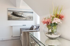Ein modernes Wohnzimmer mit einem Glastisch, einer Blumenvase, einer Couch und einem Gemälde eines Bootes an der Wand.