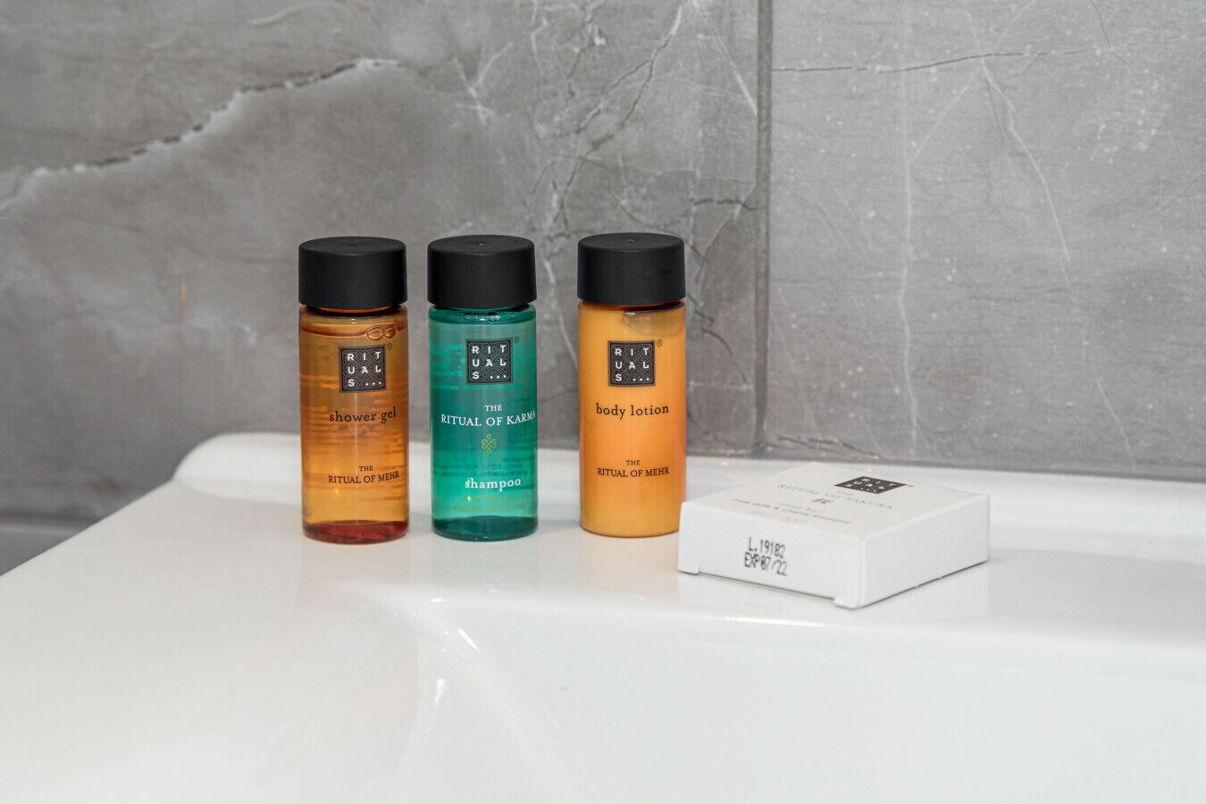 Drei Flaschen Rituals Duschgel, Shampoo und Körperlotion auf einer weißen Oberfläche mit einem grauen strukturierten Hintergrund.